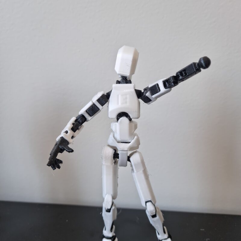 Boneco articulado branco e preto de pé, com o braço estendido, sobre fundo claro
