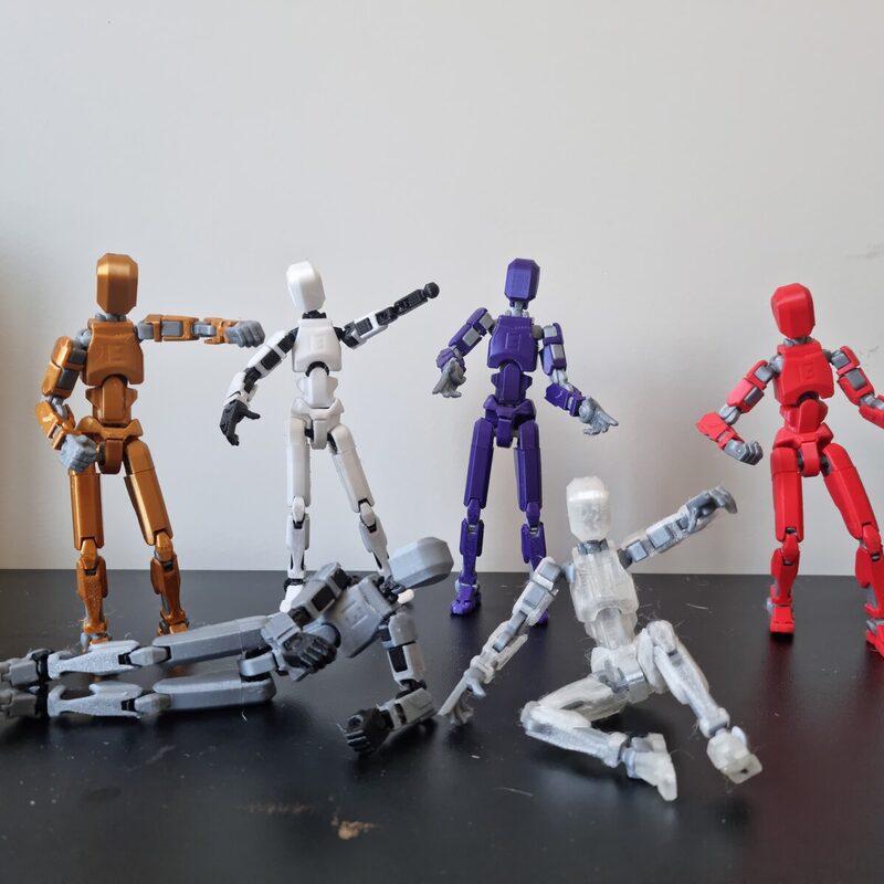 Quatro bonecos articulados impressos em 3D, nas cores laranja, branco, roxo e vermelho, sobre mesa escura