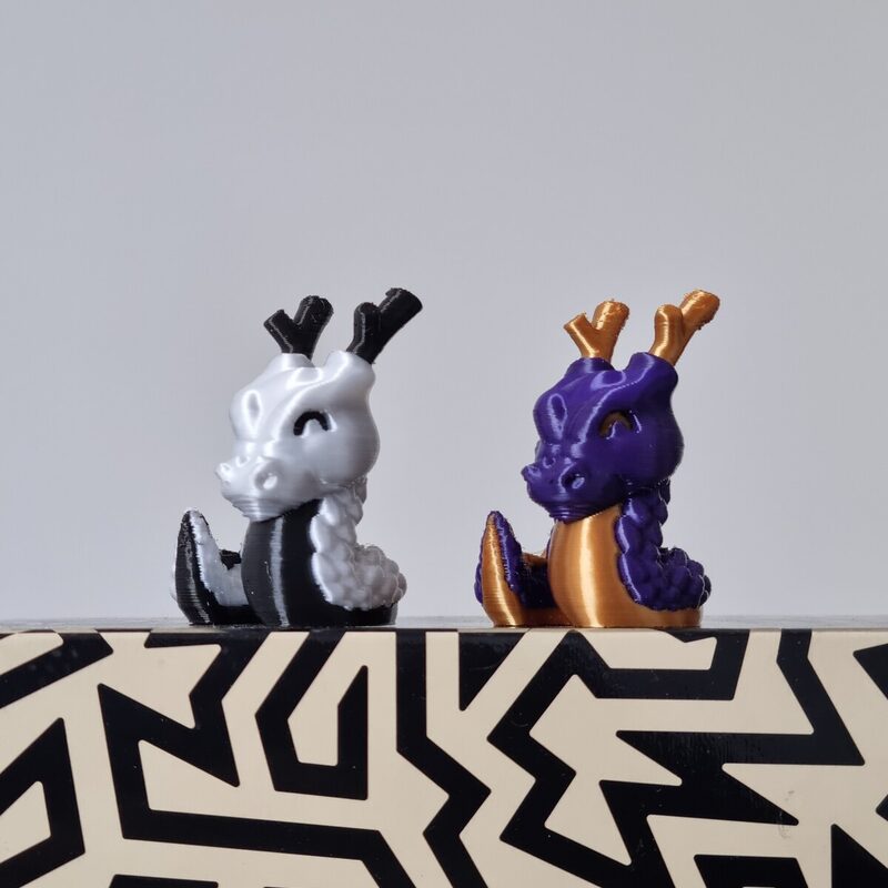 Dois dragões impressos em 3D, um branco e preto e outro roxo e dourado, lado a lado