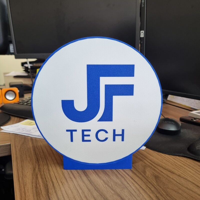 Luminária circular impressa em 3D com o logotipo azul da JF Tech, acesa sobre uma mesa de escritório