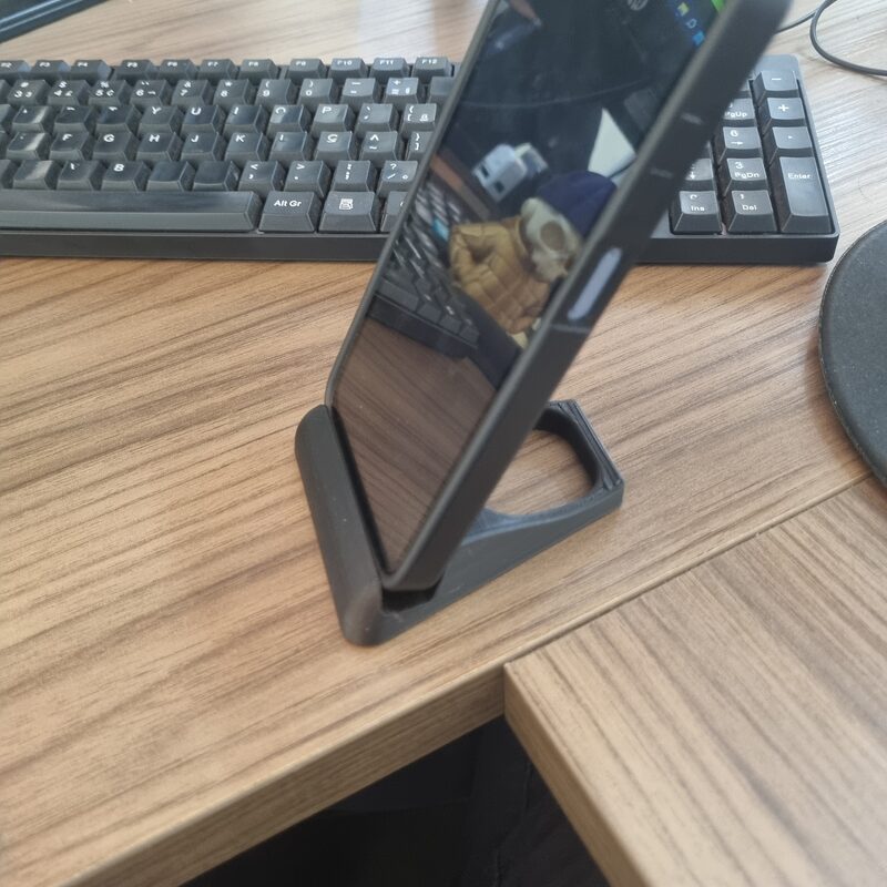 Celular apoiado em um suporte impresso em 3D em filamento cinza, sobre mesa de madeira